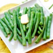 Boiled green beans are served on a white plate, topped with butter.