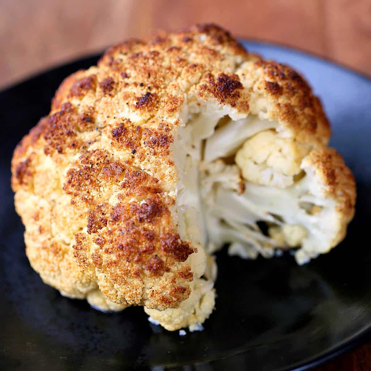 Roasted whole cauliflower is served on a dark plate.