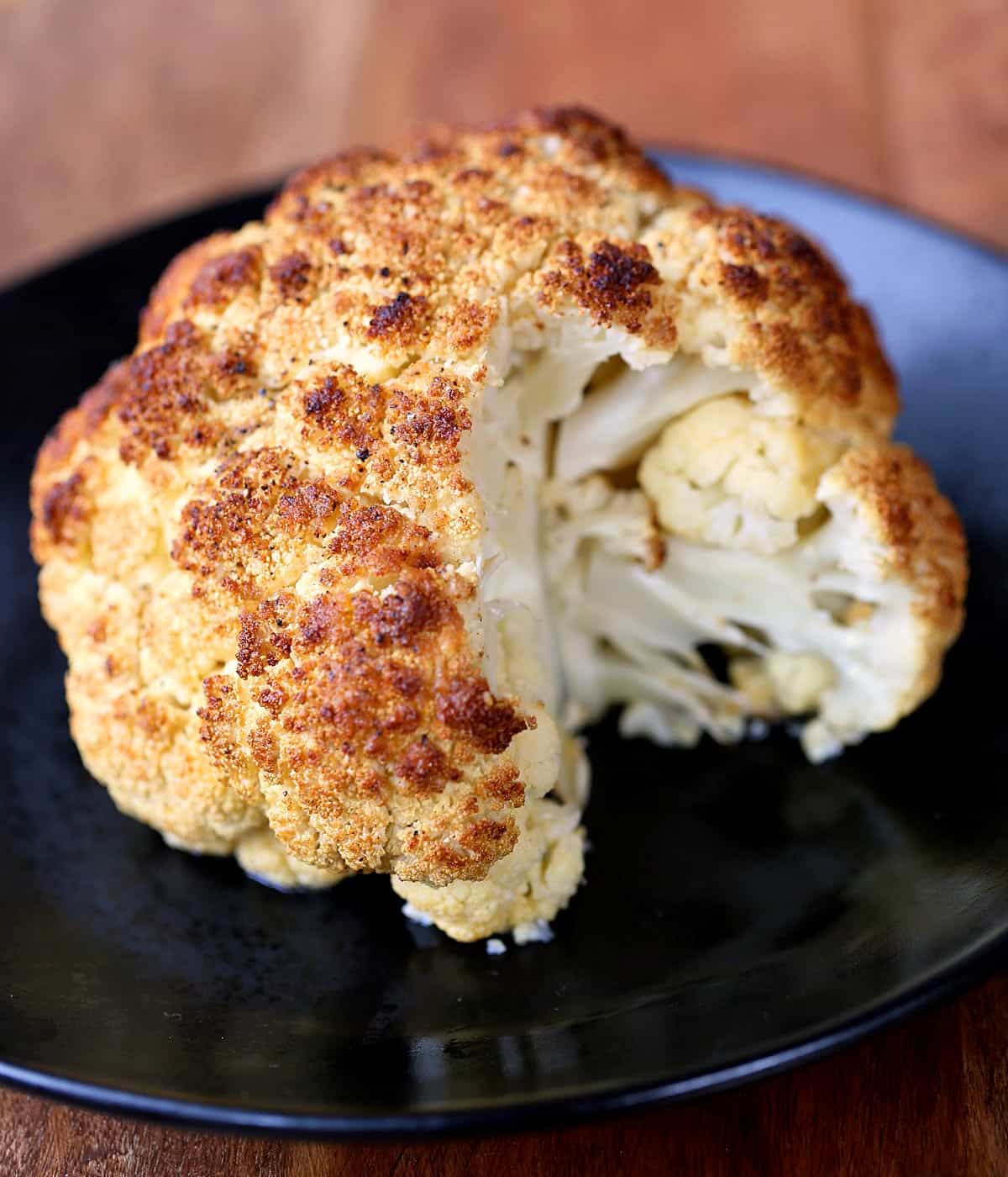 Whole roasted cauliflower is served on a dark plate. 