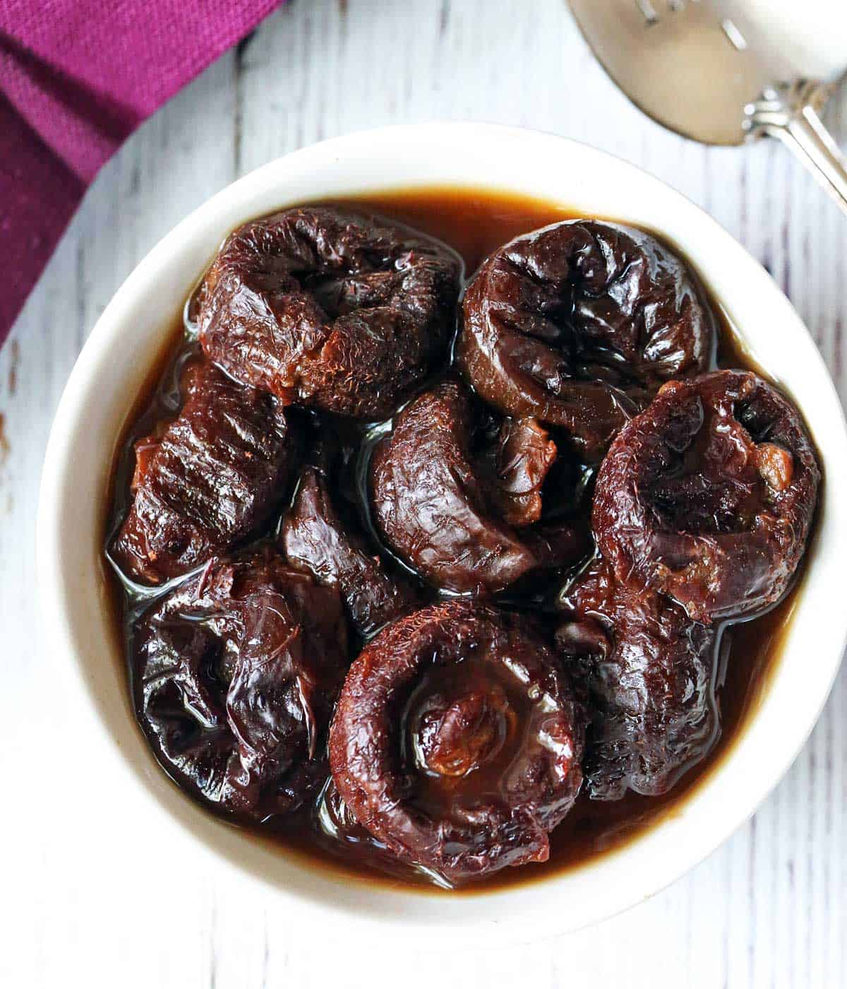 Stewed prunes are served in a white bowl with a napkin and a spoon. 