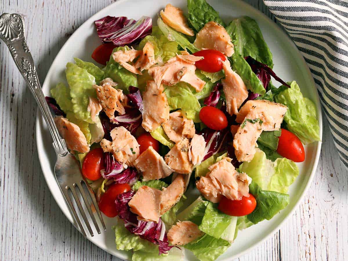 Using up leftover baked salmon in a salad. The salad contains lettuce and tomatoes and served on a white plate.