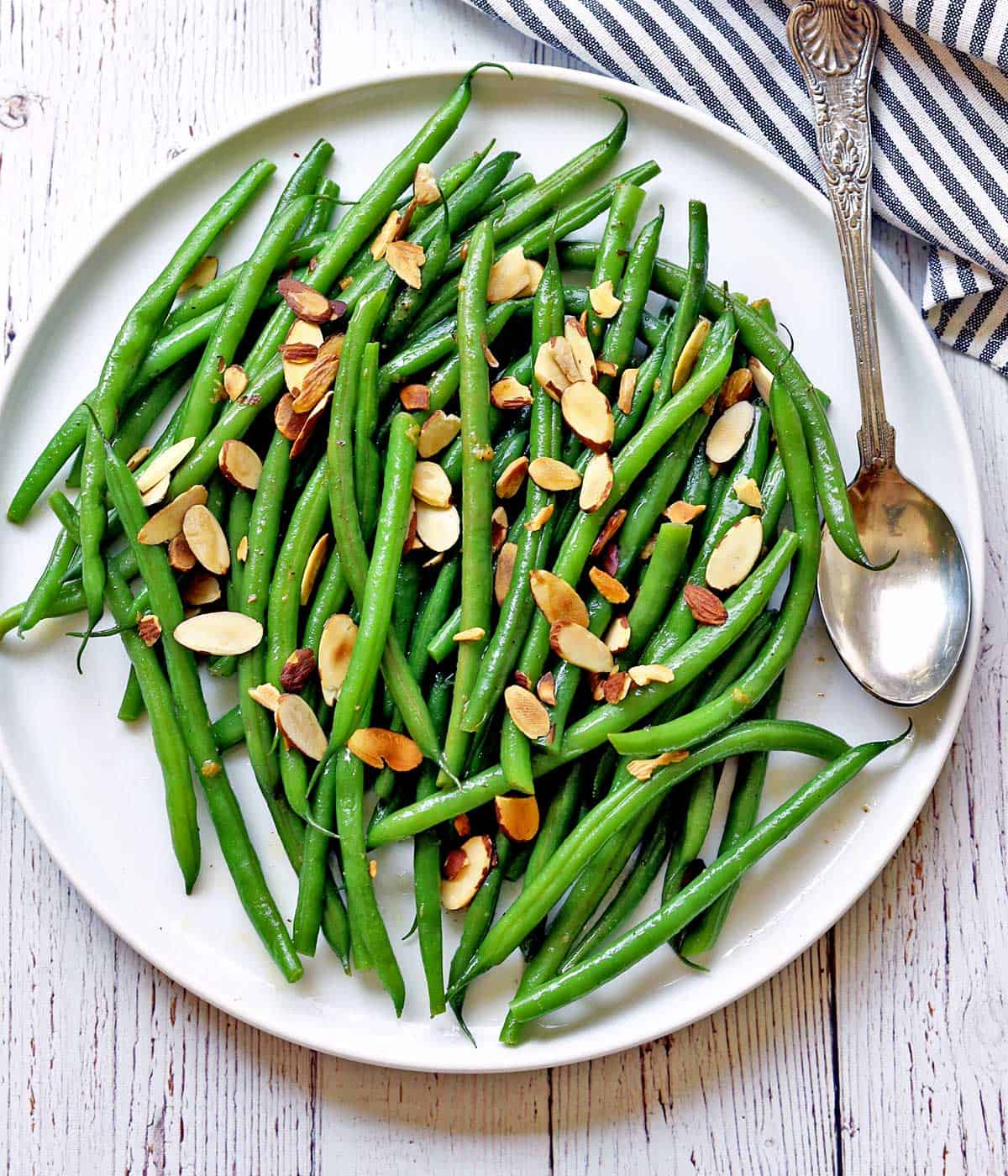 Green beans almondine are served on a white plate with a serving spoon. 