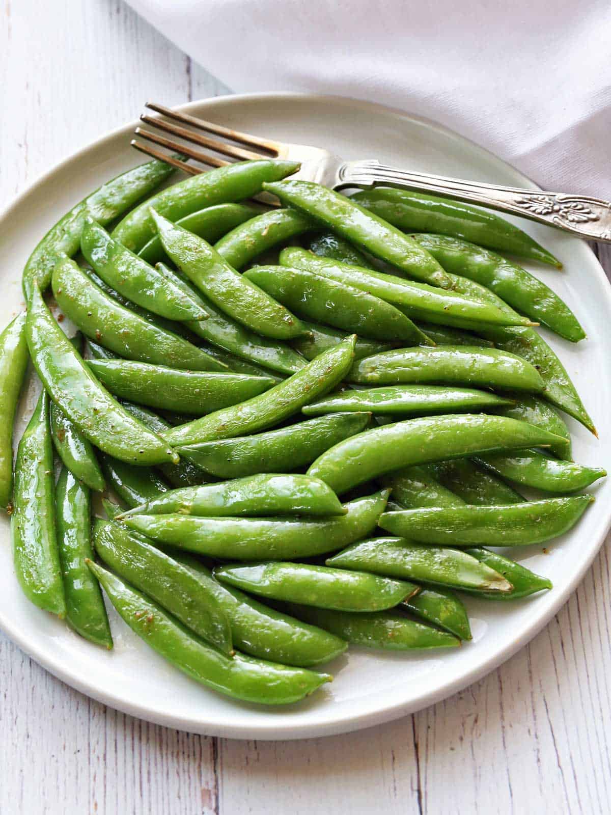 Sauteed sugar snap peas are served on a white plate with a silver pork. 