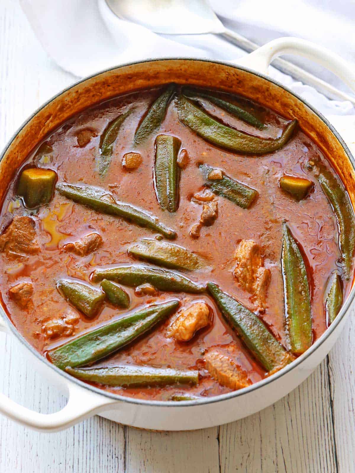 Okra stew is served in a white saucepan with a serving spoon. 