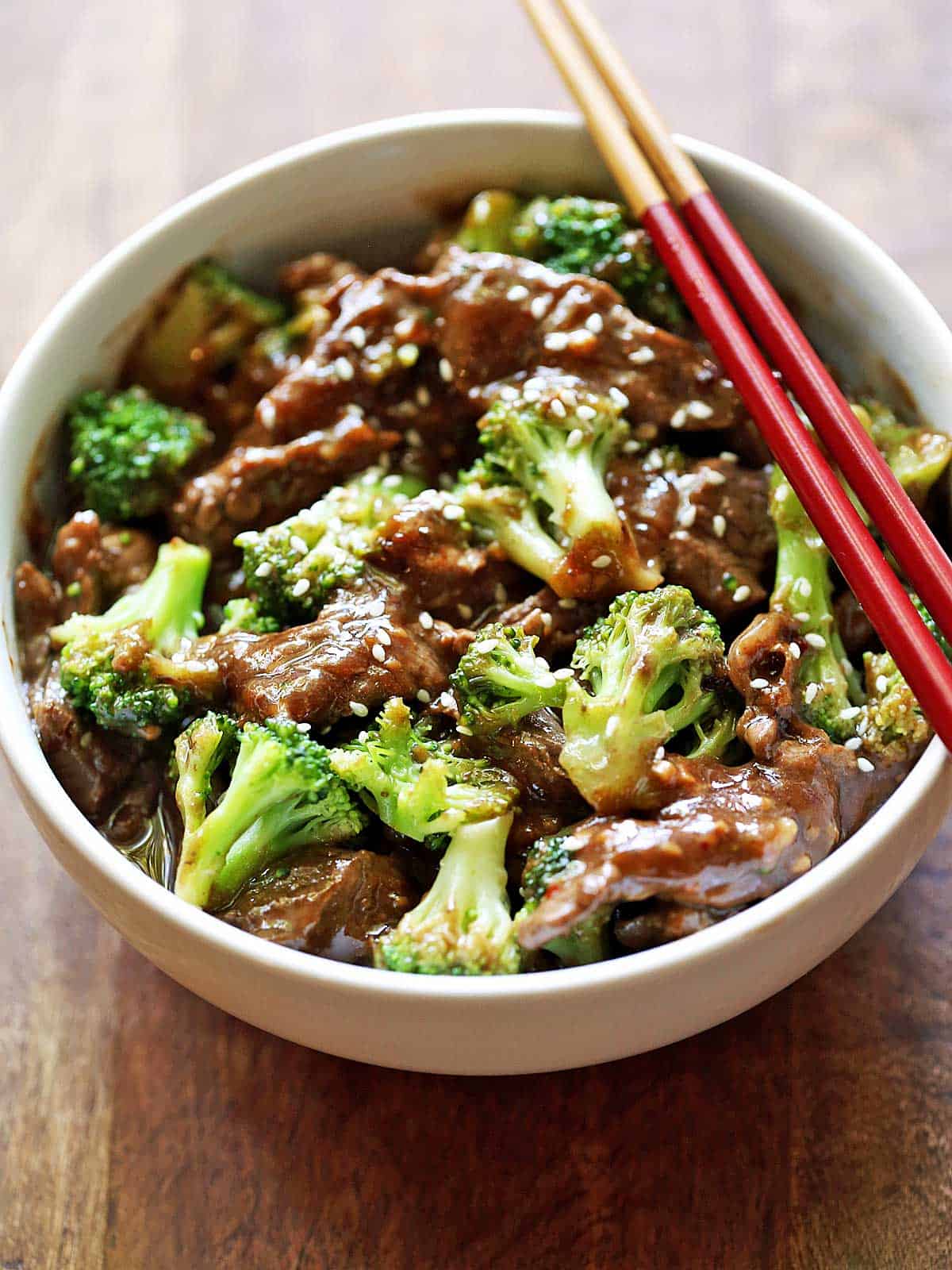 Beef and broccoli is served in a white bowl with red chopsticks. 