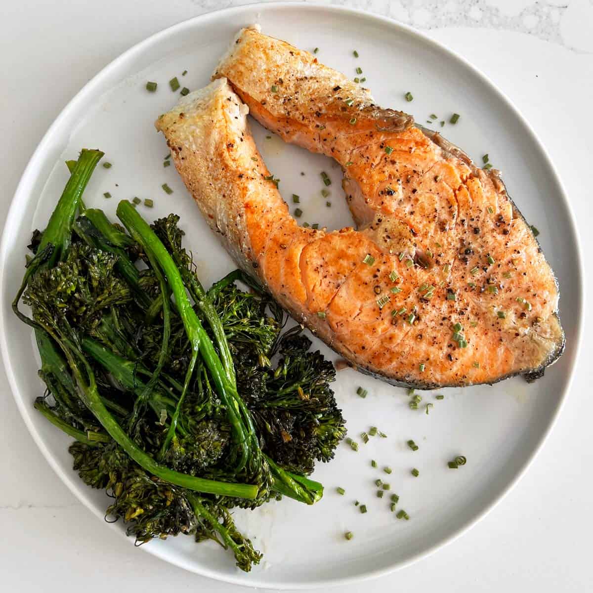 A salmon steak is served on a white plate with broccolini.