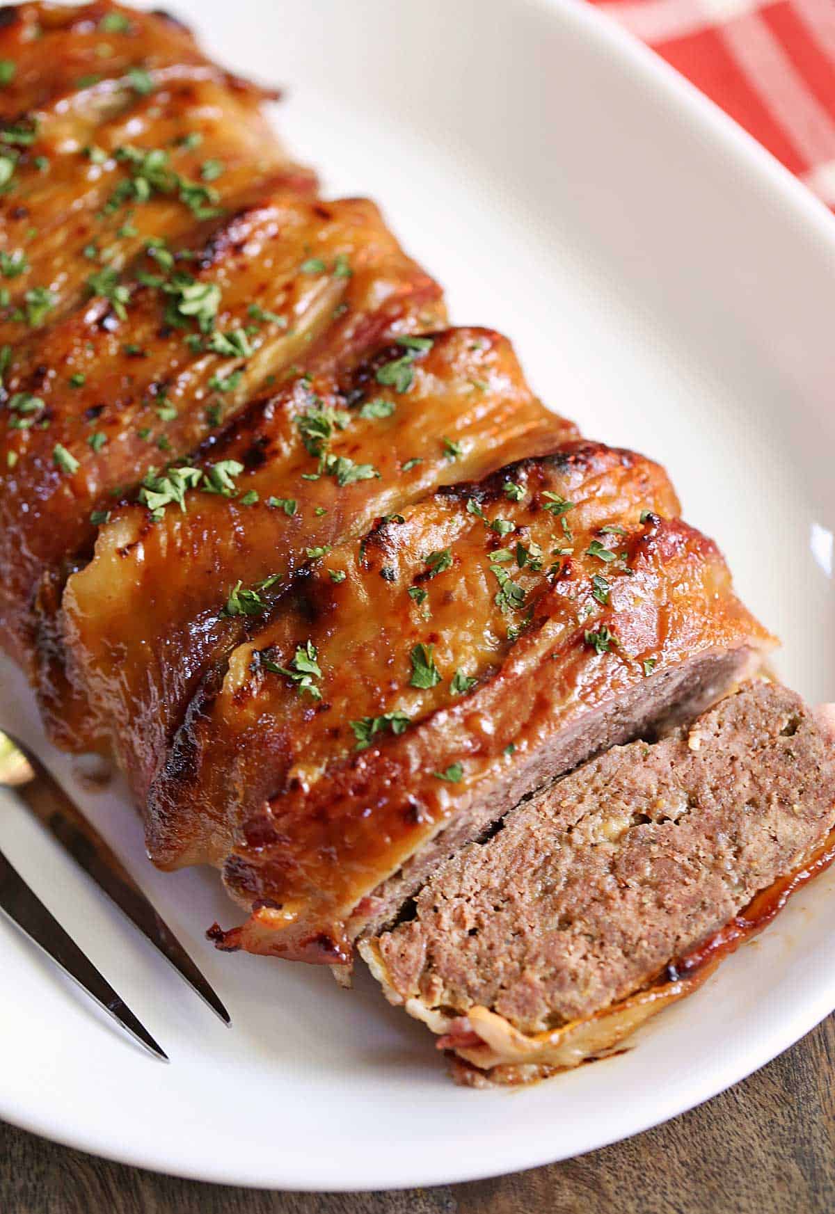 Bacon wrapped meatloaf is served on a white platter with a serving fork.