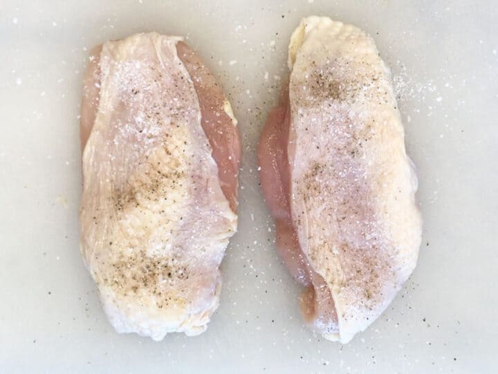 Two skin-on chicken breasts are seasoned with salt and pepper.