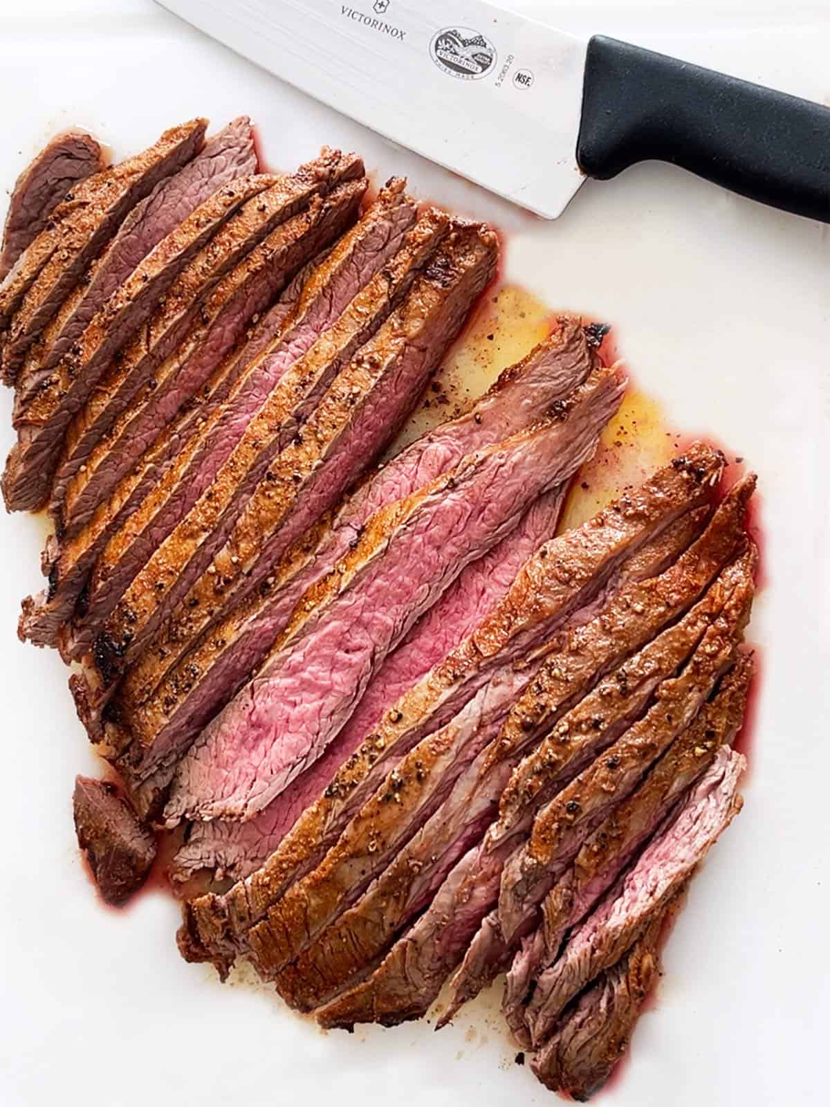 A sliced flank steak is served on a white cutting board. 