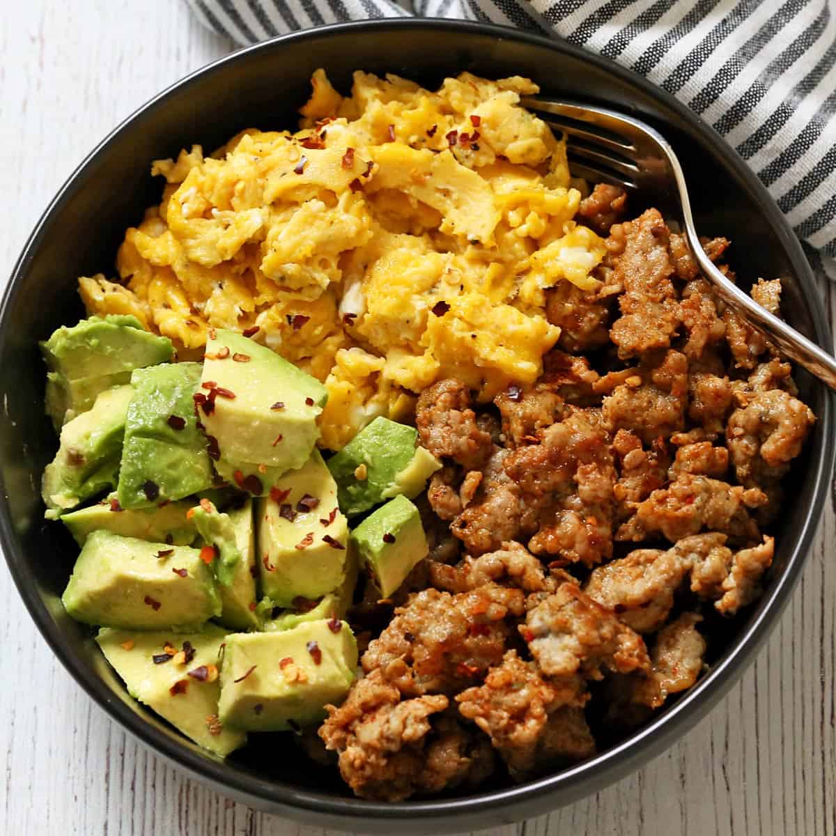 A breakfast bowl with sausage, scrambled eggs, and avocado is served with a napkin and a fork.