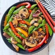 A vegetable stir-fry is served in a black bowl with red chopsticks.