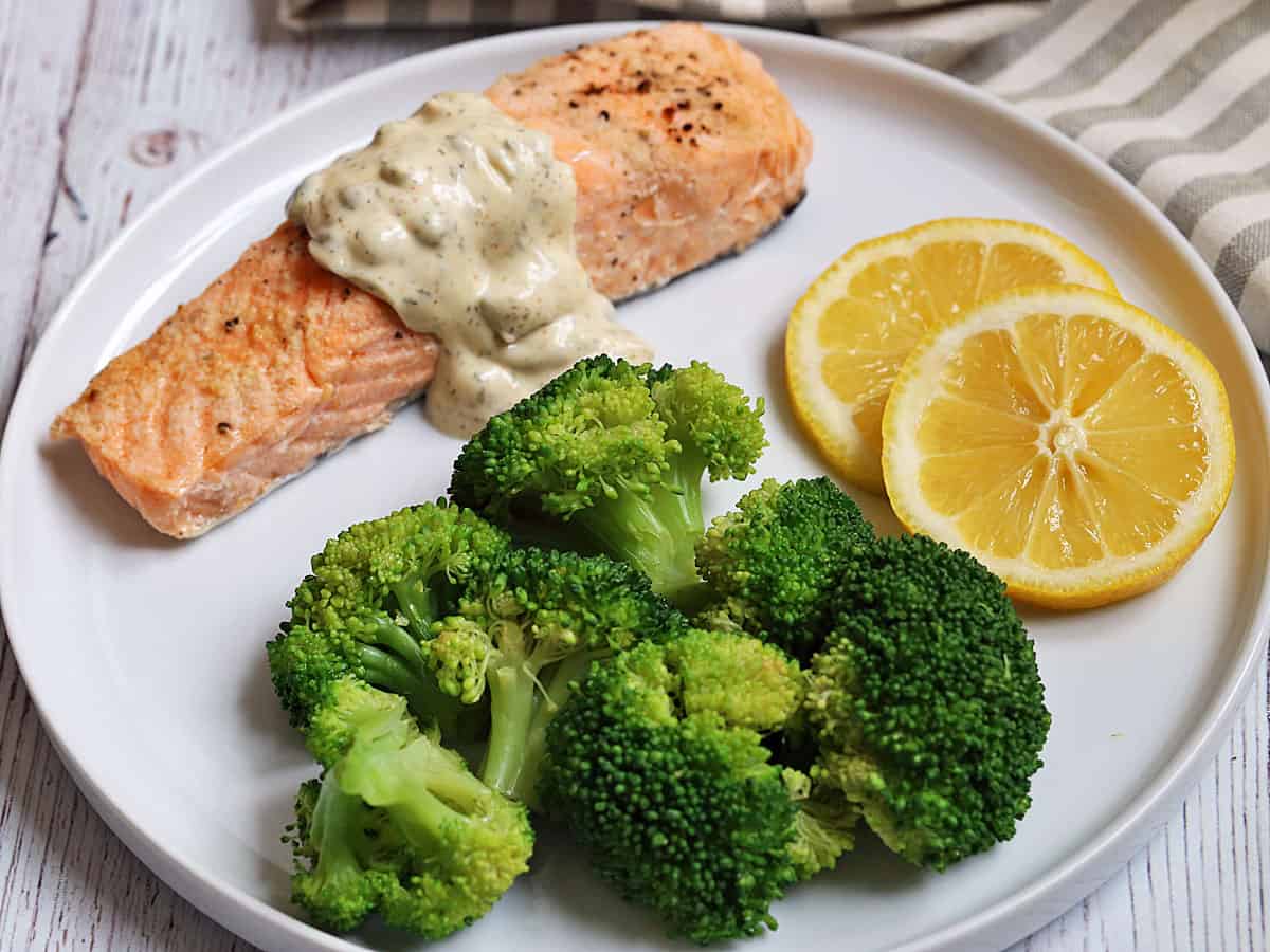 Poached salmon is served with tartar sauce, steamed broccoli, and lemon slices.
