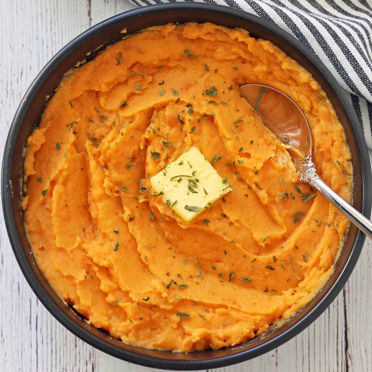 Mashed sweet potatoes are served in a dark bowl with a serving spoon.