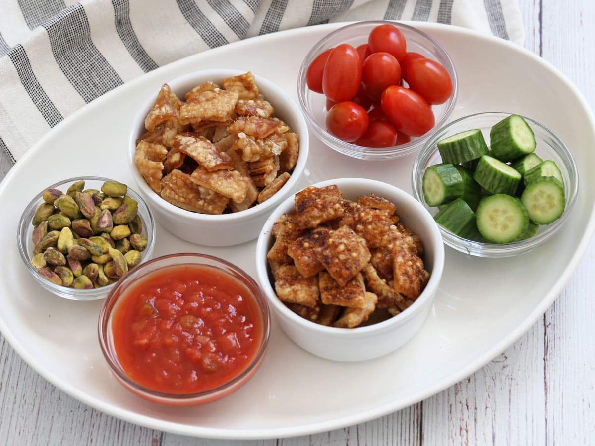 A snack platter with pork rinds, nuts, salsa, and veggies.