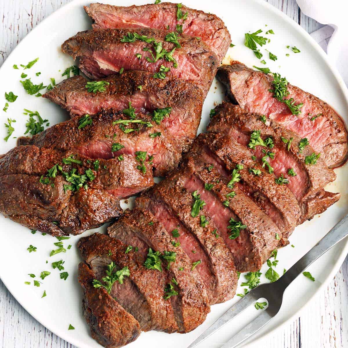 A sliced flat iron steak is served on a white plate with a serving fork.