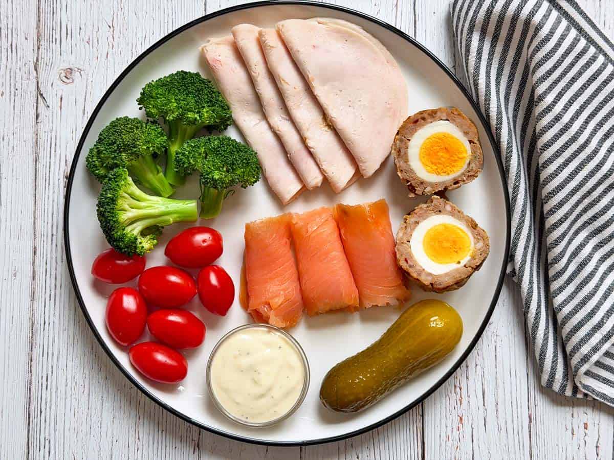 A snack plate with Scotch eggs, smoked salmon, roasted chicken lunchmeat, ranch dressing, veggies, and pickles.