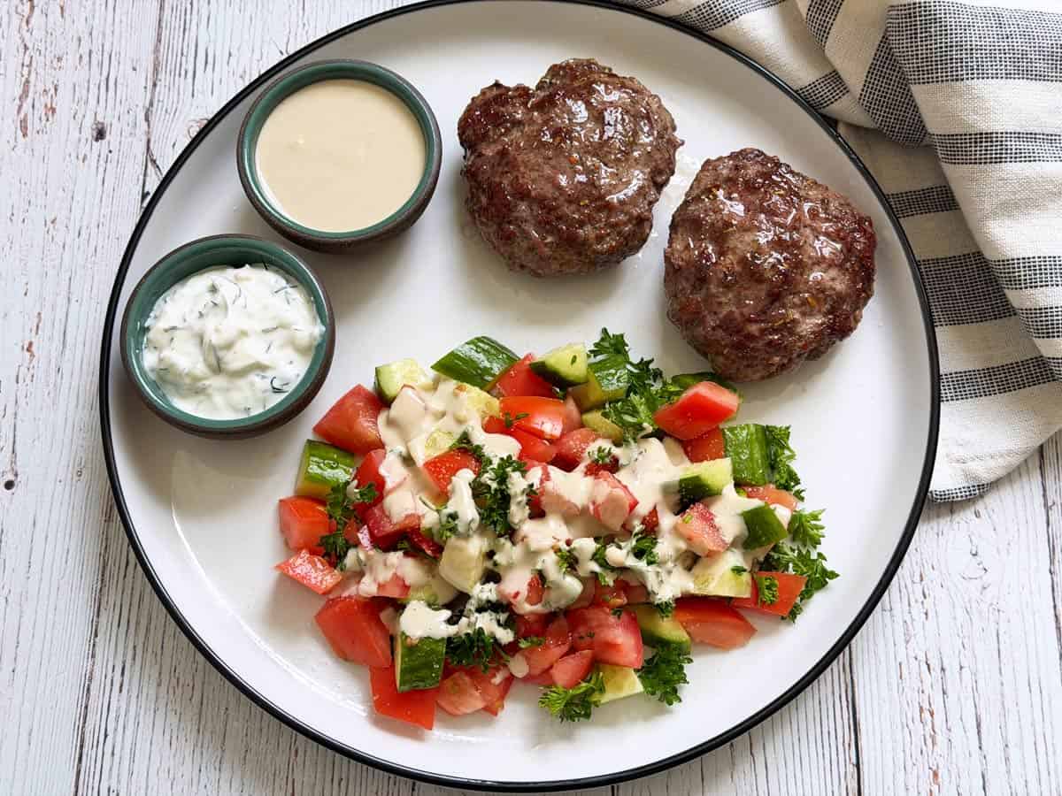 Tahini sauce is drizzled over Israeli salad. Also on the plate: lamb burgers and tzatziki.