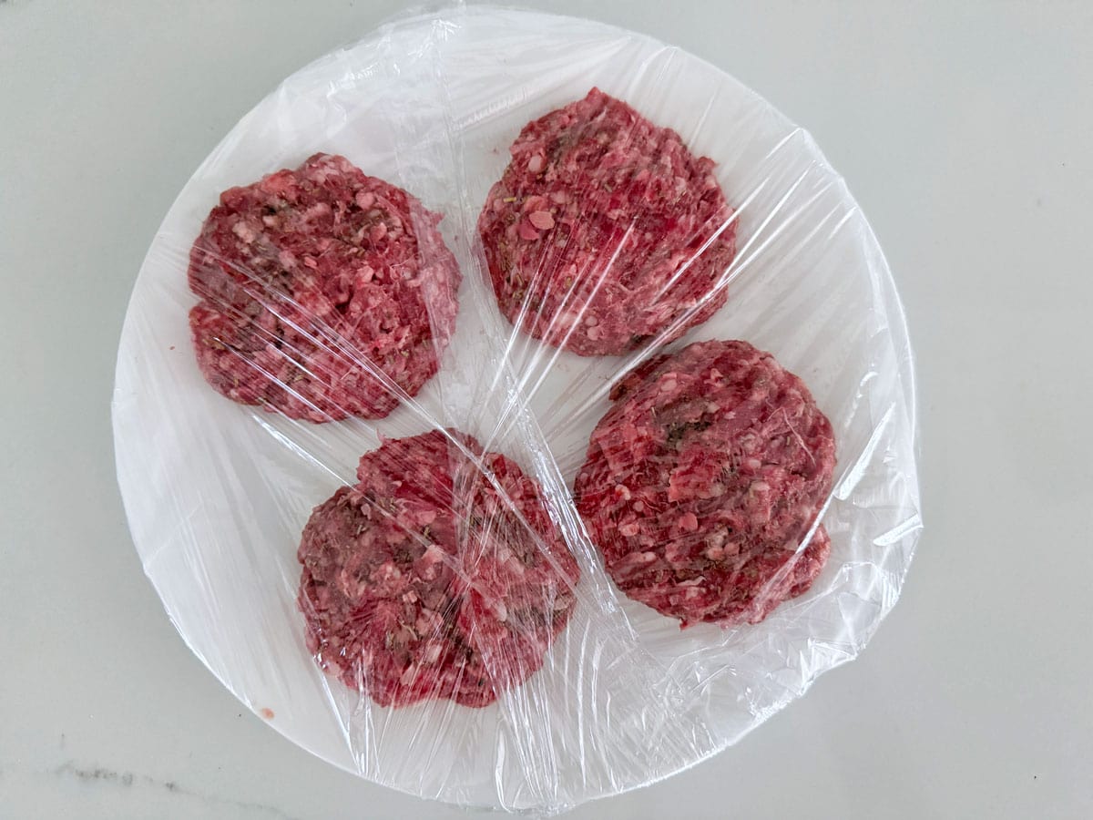 Lamb burgers on a plate, covered with plastic wrap.