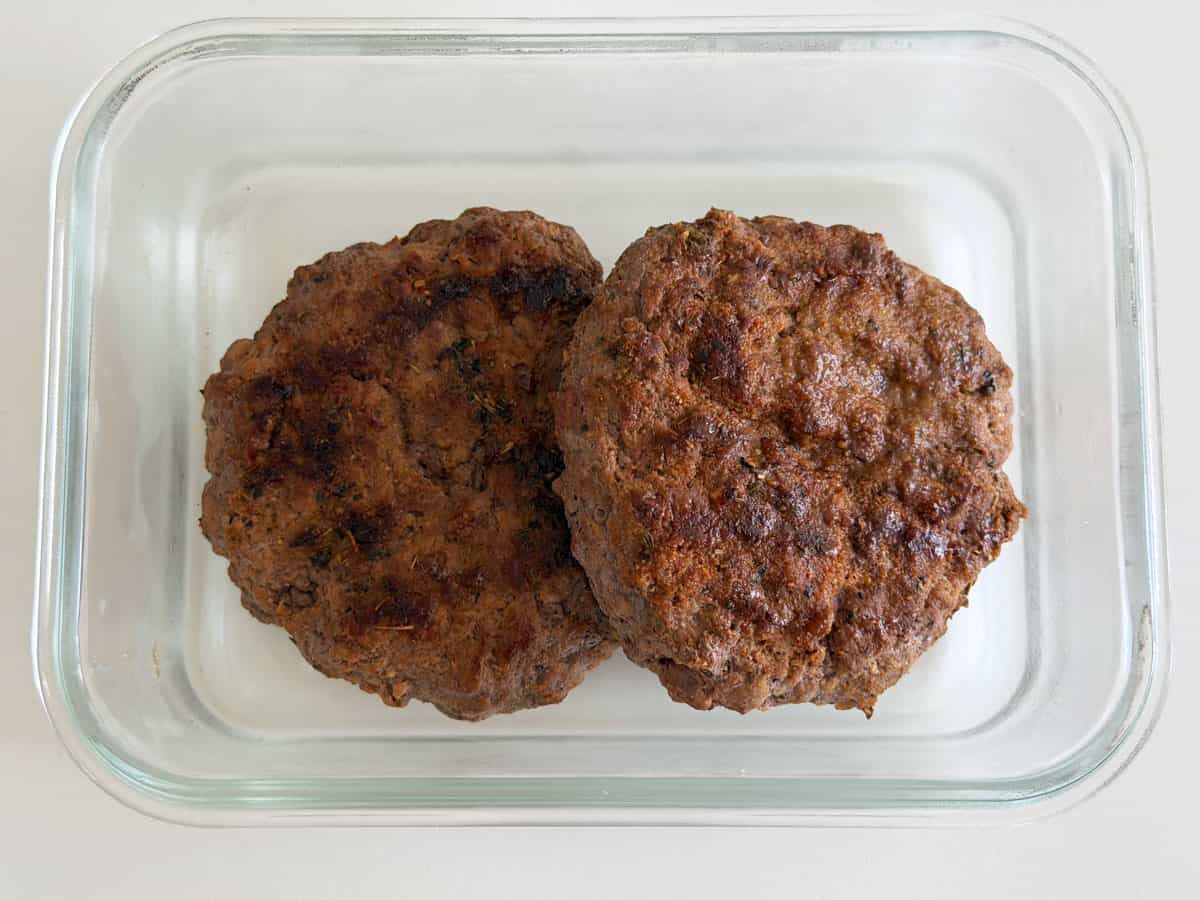 Two leftover grilled burgers are stored in a glass container.